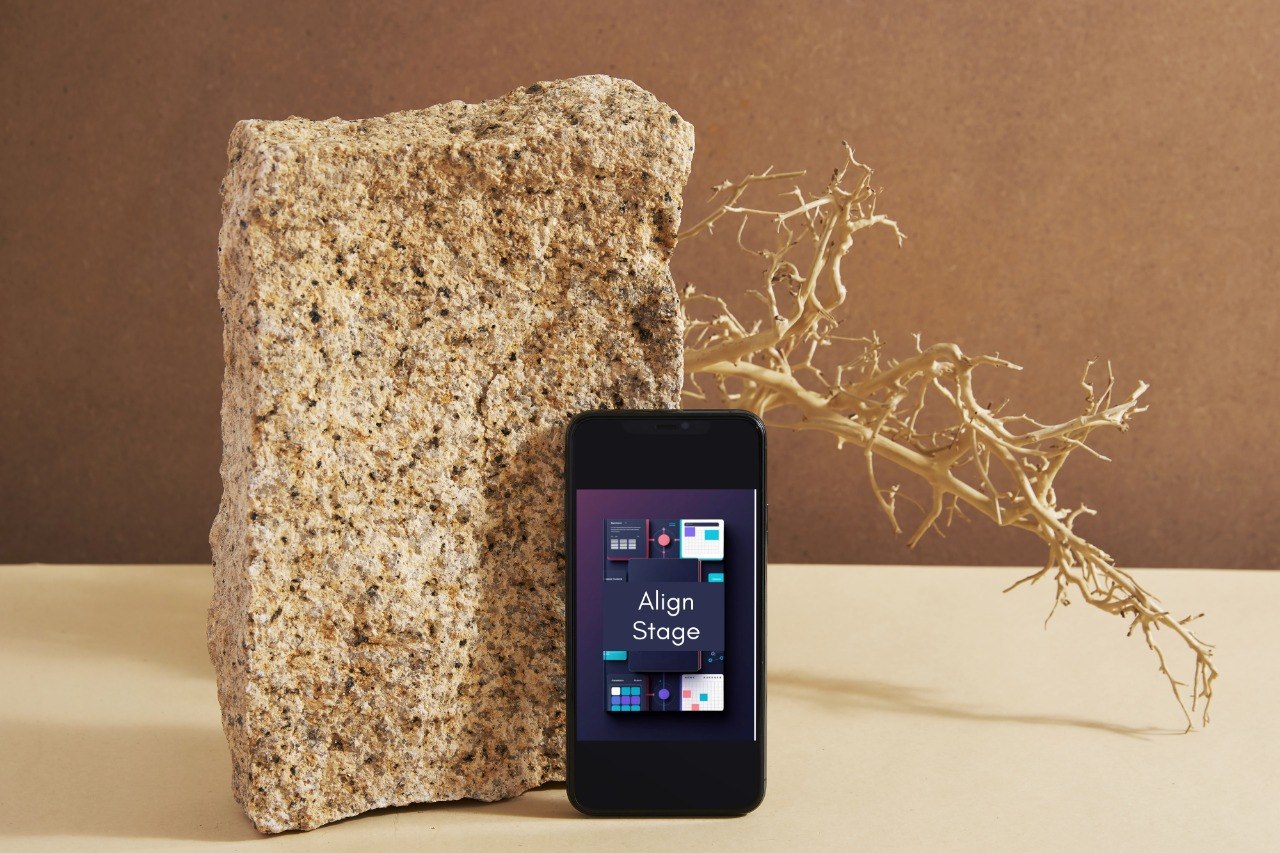 A black smartphone with its screen on, displaying a Windows-style interface and the text 'Align Stage,' placed in front of a large rough stone on a beige tabletop against a brown backdrop, with a dried branch behind it.