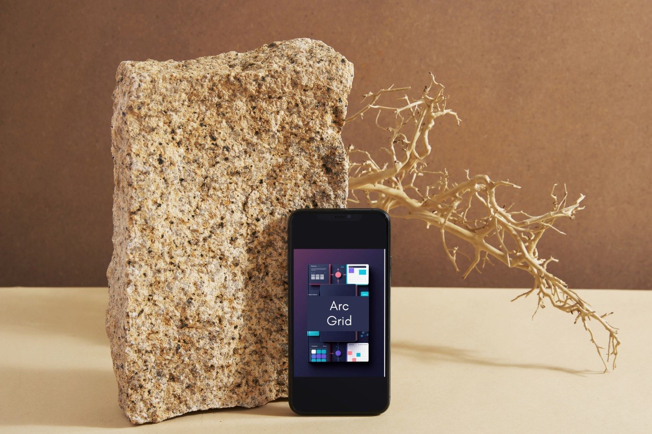 A black smartphone displaying a Windows-style desktop interface labeled 'Mode Grid' stands in front of a large rough stone on a beige surface, with a dried branch against a brown backdrop.