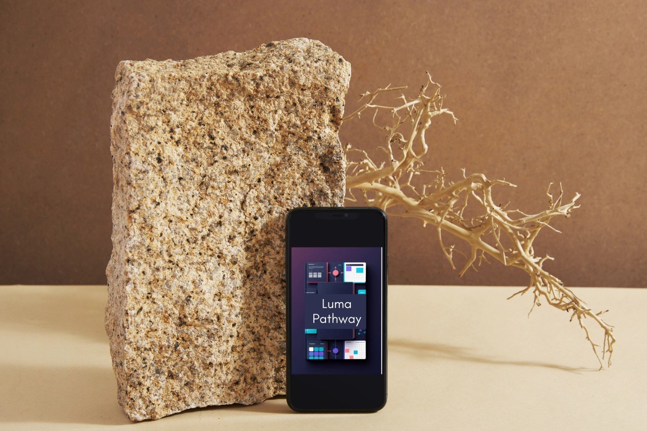 A black smartphone displaying the Luma Pathway app stands upright next to a large rough stone on a beige surface, with a dried branch against a brown backdrop.