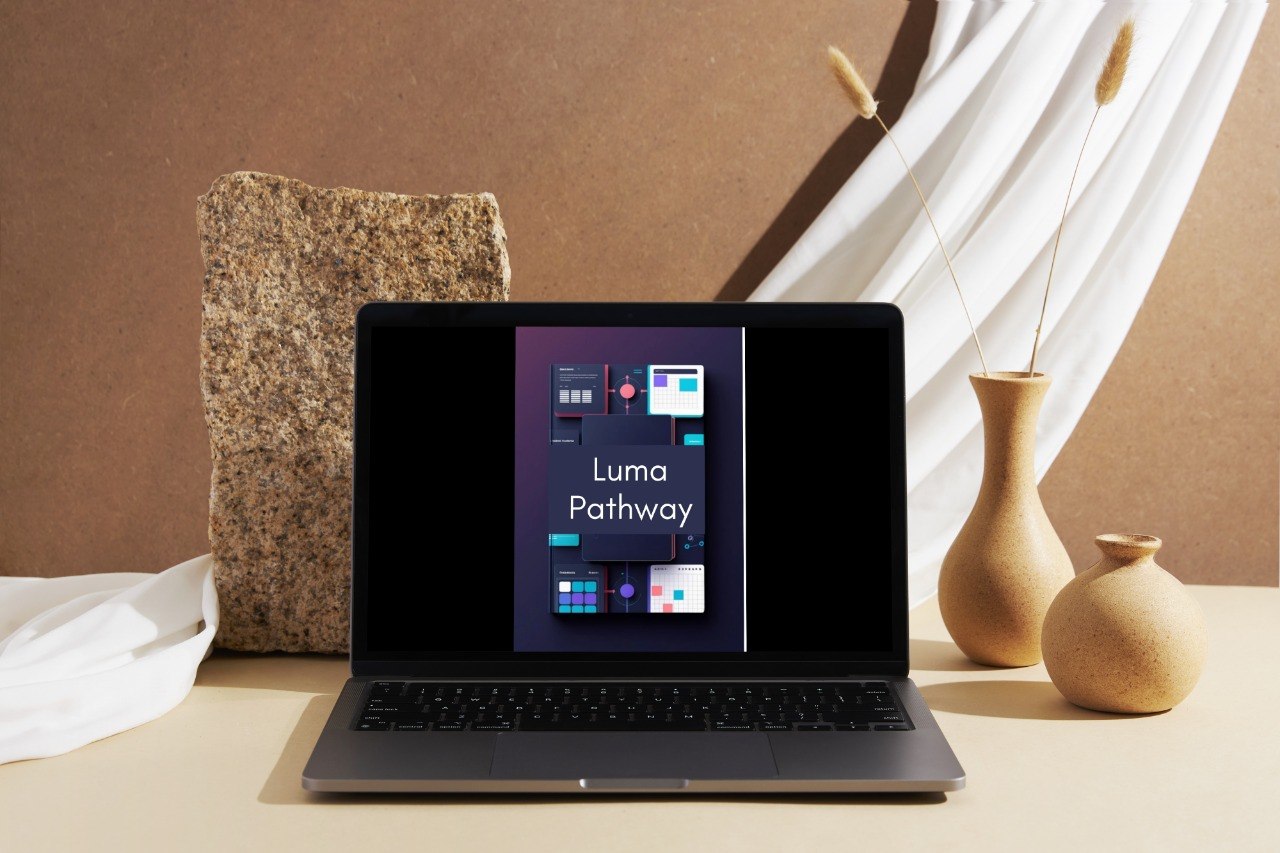 A dark gray laptop sits open on a beige tabletop, displaying the Luma Pathway interface on its screen. The setup is styled with a textured stone block, draped white fabric, and two beige ceramic vases against a warm brown background.